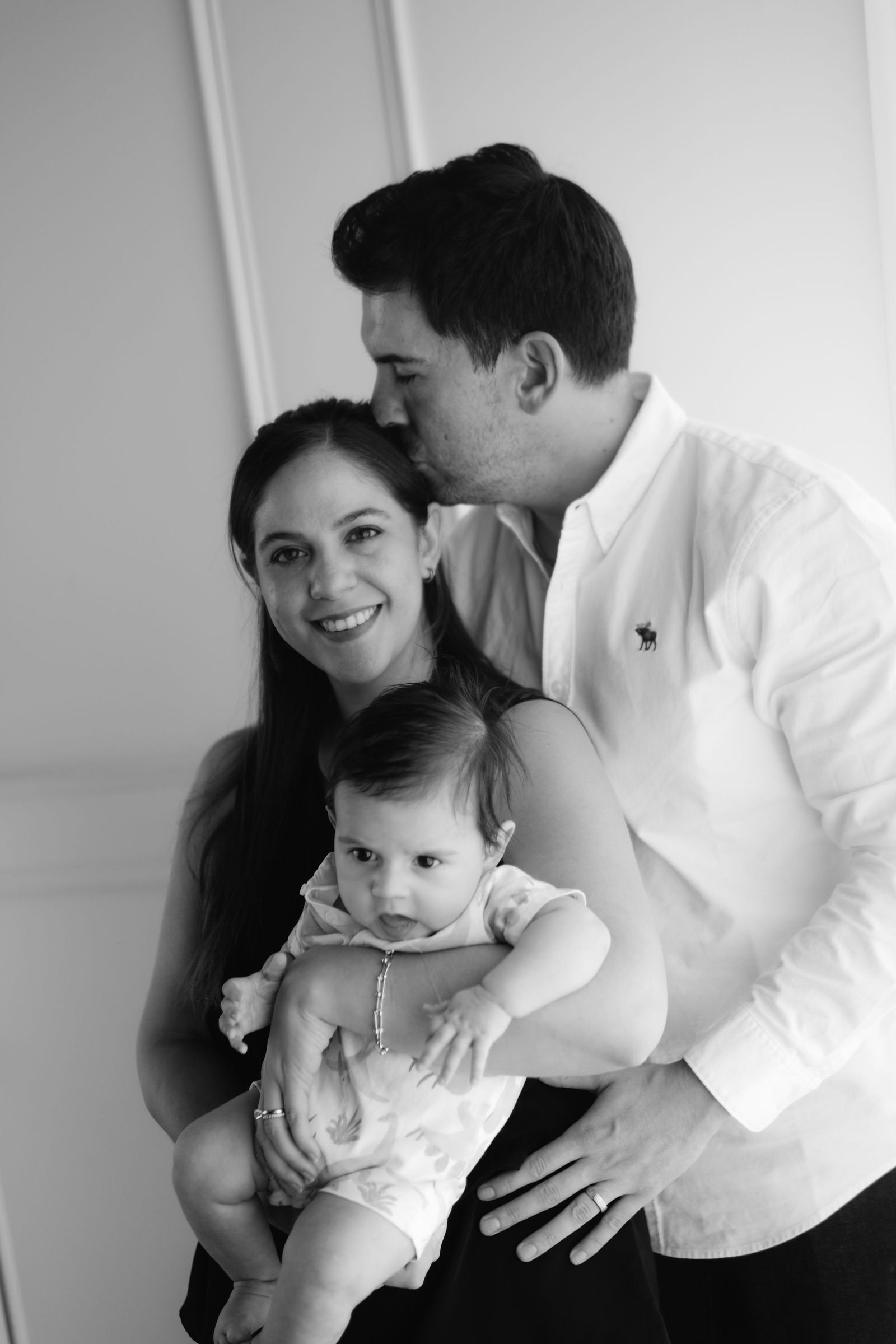 En blanco y negro, un papá besa en la frente a la mamá, que sostiene a su bebé.