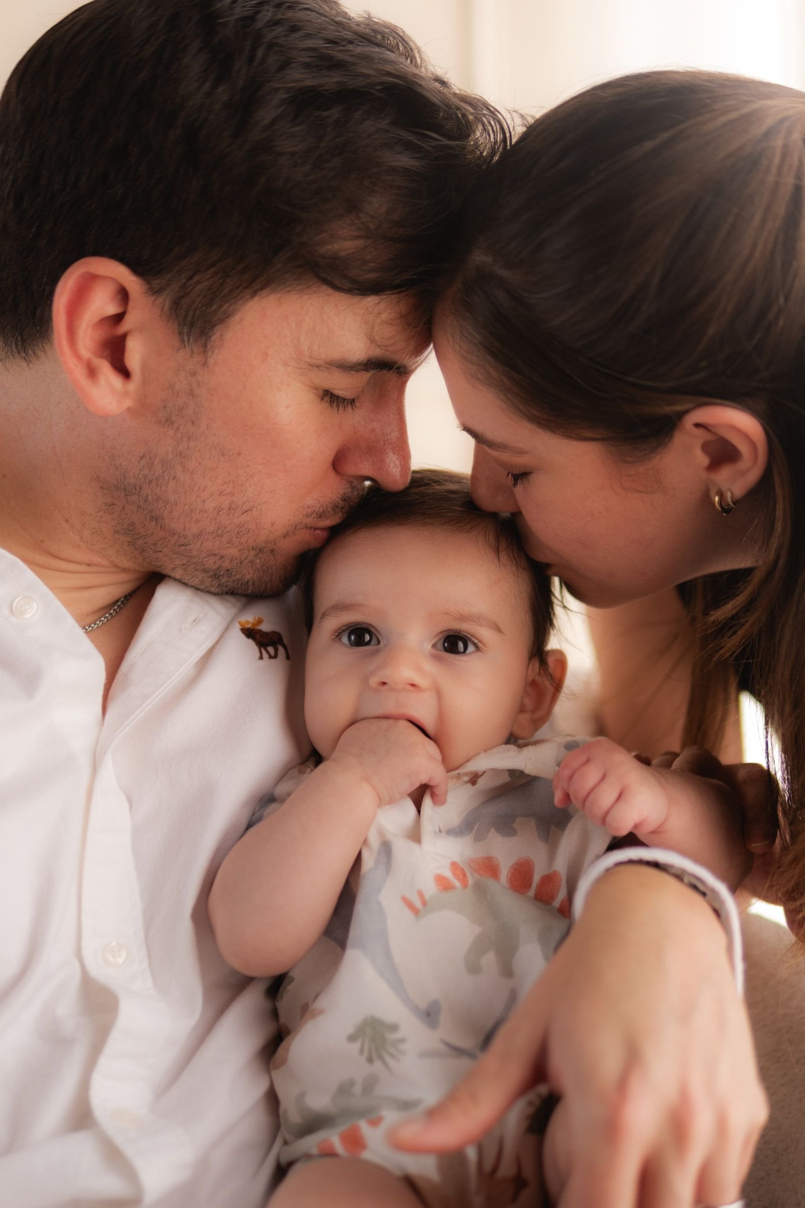 Papá y mamá besan a su bebé, que se lleva la mano a la boca.
