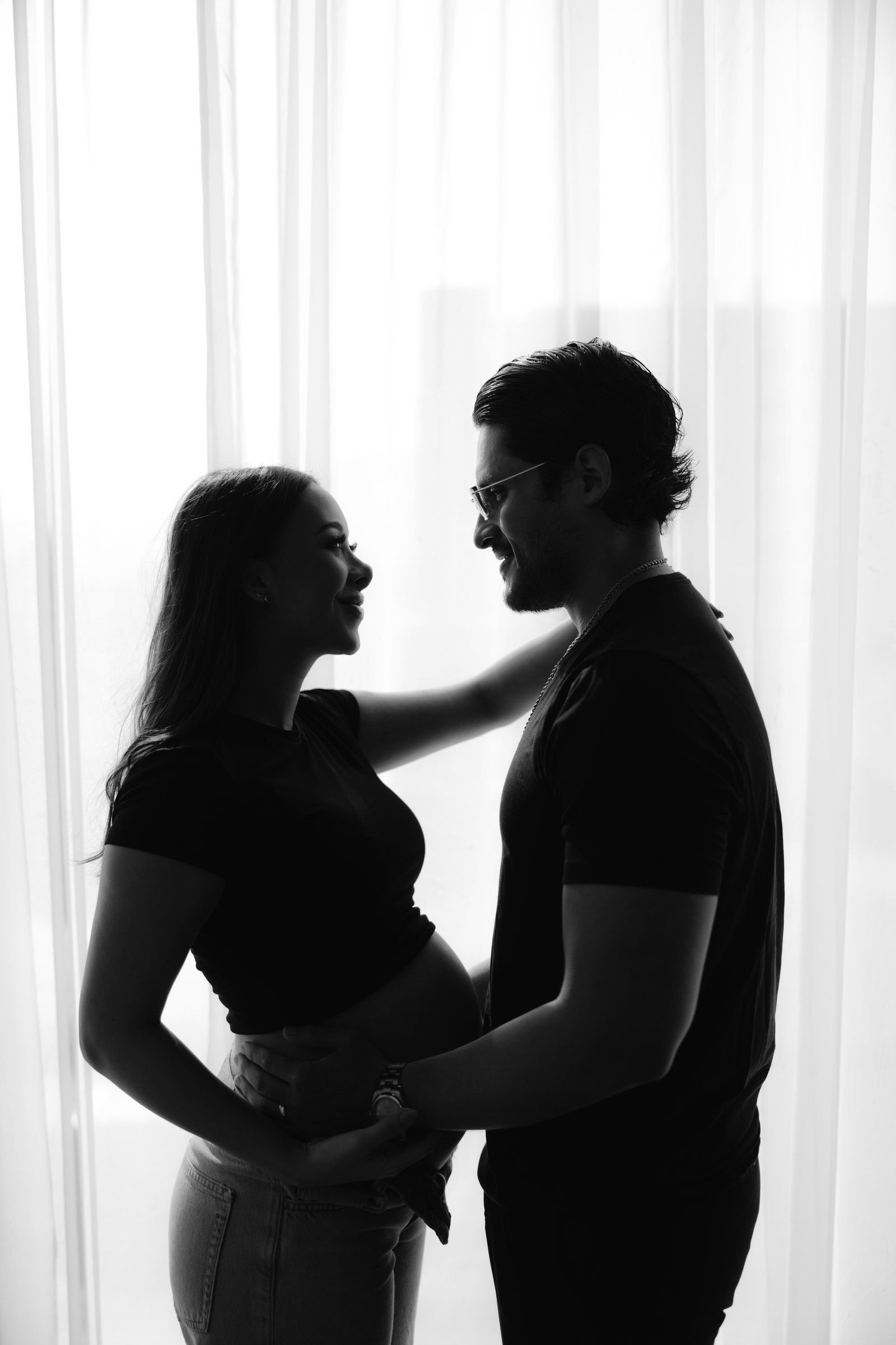 Silueta de una pareja que espera un bebé, abrazados frente a una ventana luminosa.
