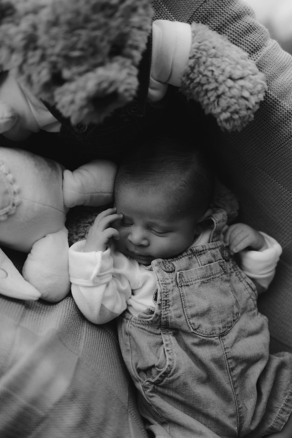 En blanco y negro, un bebé recién nacido duerme sobre una cobija, con un oso de peluche cerca.