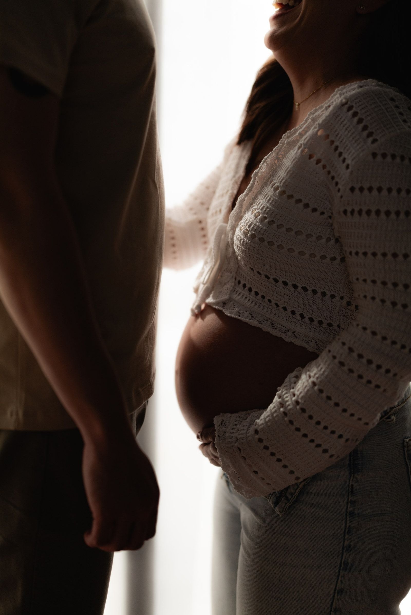 Detalle del vientre de una mujer embarazada a contraluz, con un suéter de punto blanco.