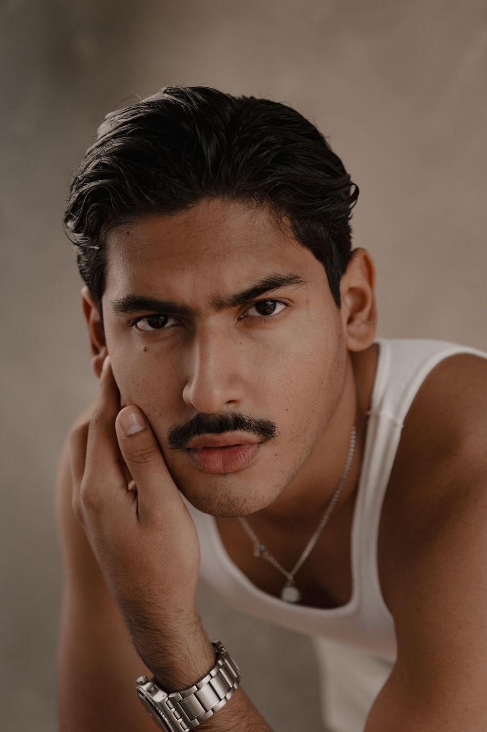 Un hombre con bigote y camiseta blanca apoya la cara en su mano y mira directo a la cámara.