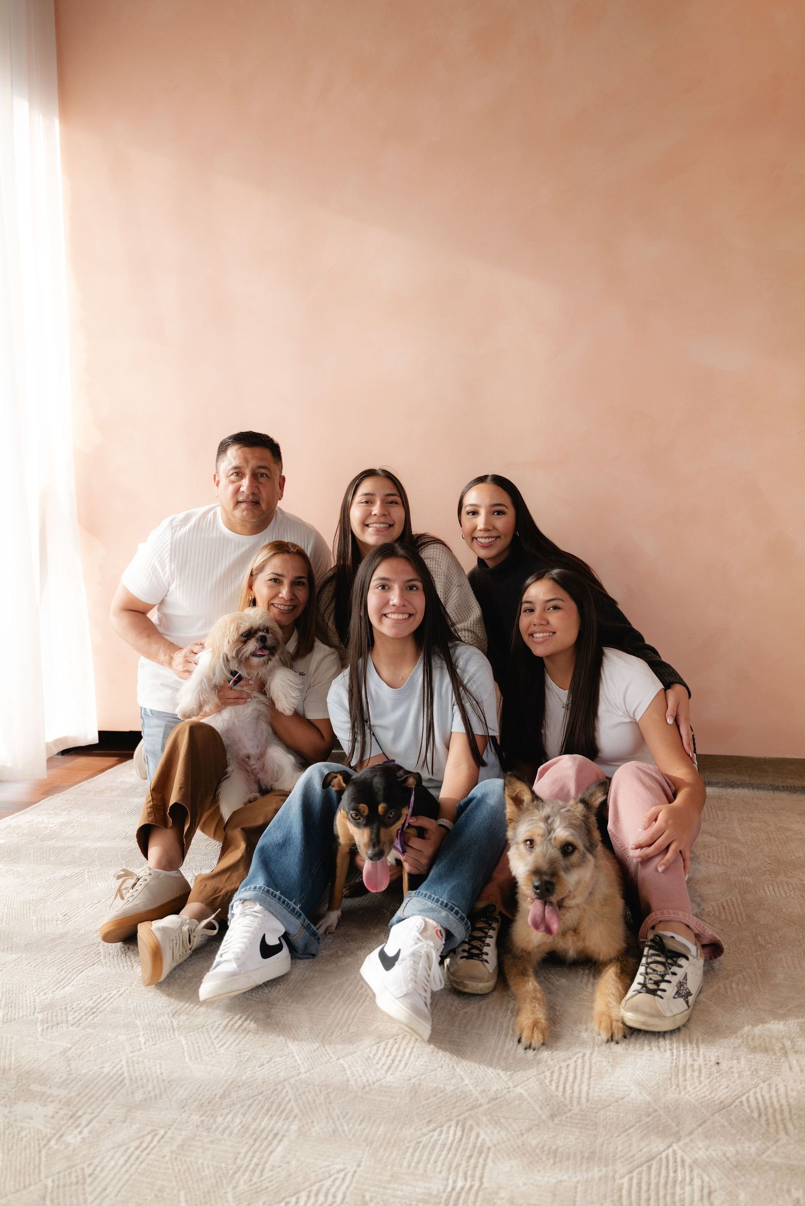 Una familia grande se sienta en el piso junto a sus perros, frente a una pared color durazno.