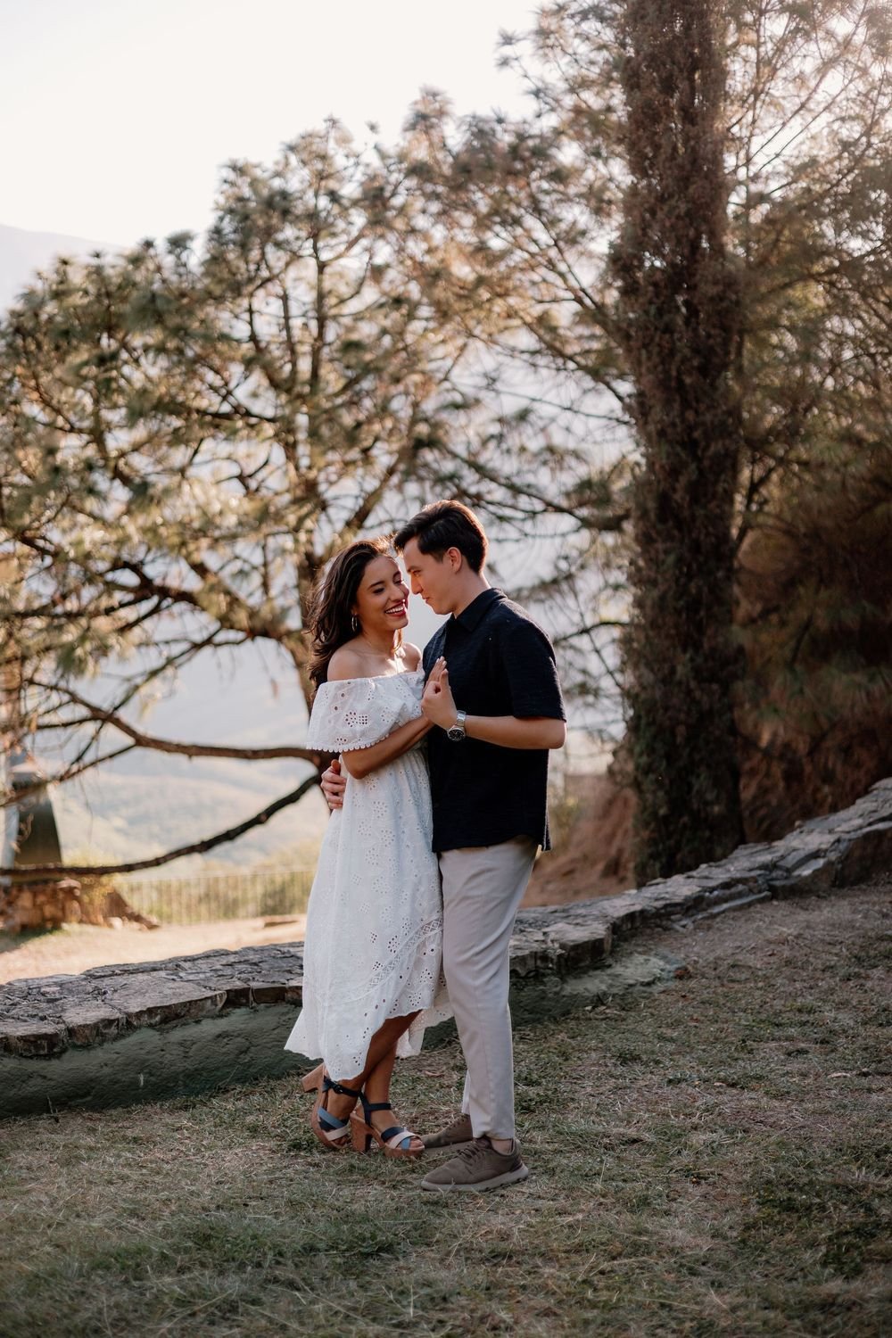 Una pareja se toma de la mano bajo un pino, al atardecer; ella lleva un vestido blanco.