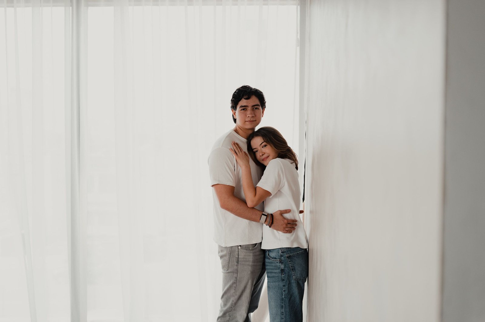 Una pareja se abraza de pie en un cuarto luminoso, junto a unas cortinas blancas.