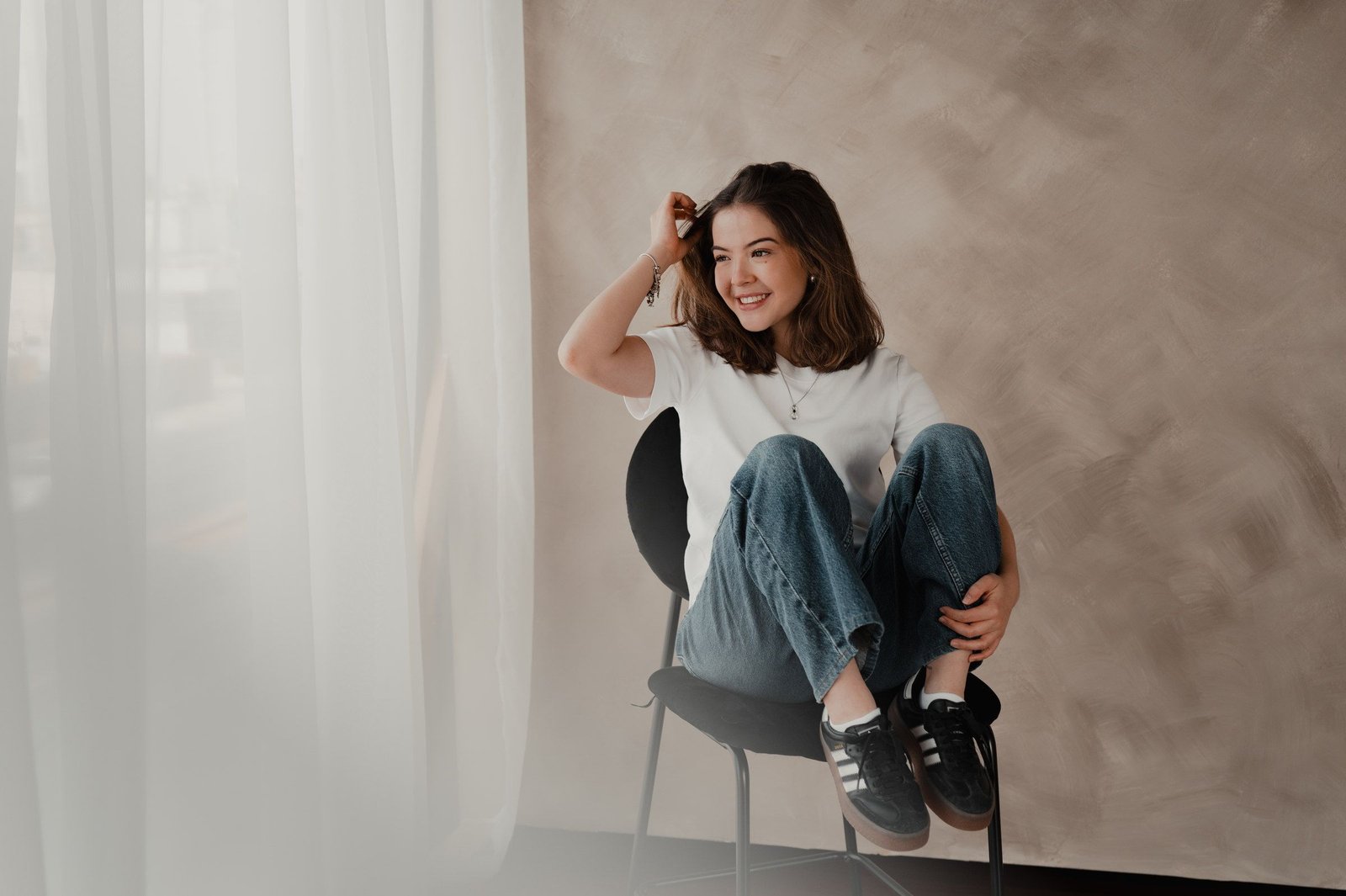 Una mujer con playera blanca y jeans se sienta en una silla negra, riendo, frente a una pared color arena.
