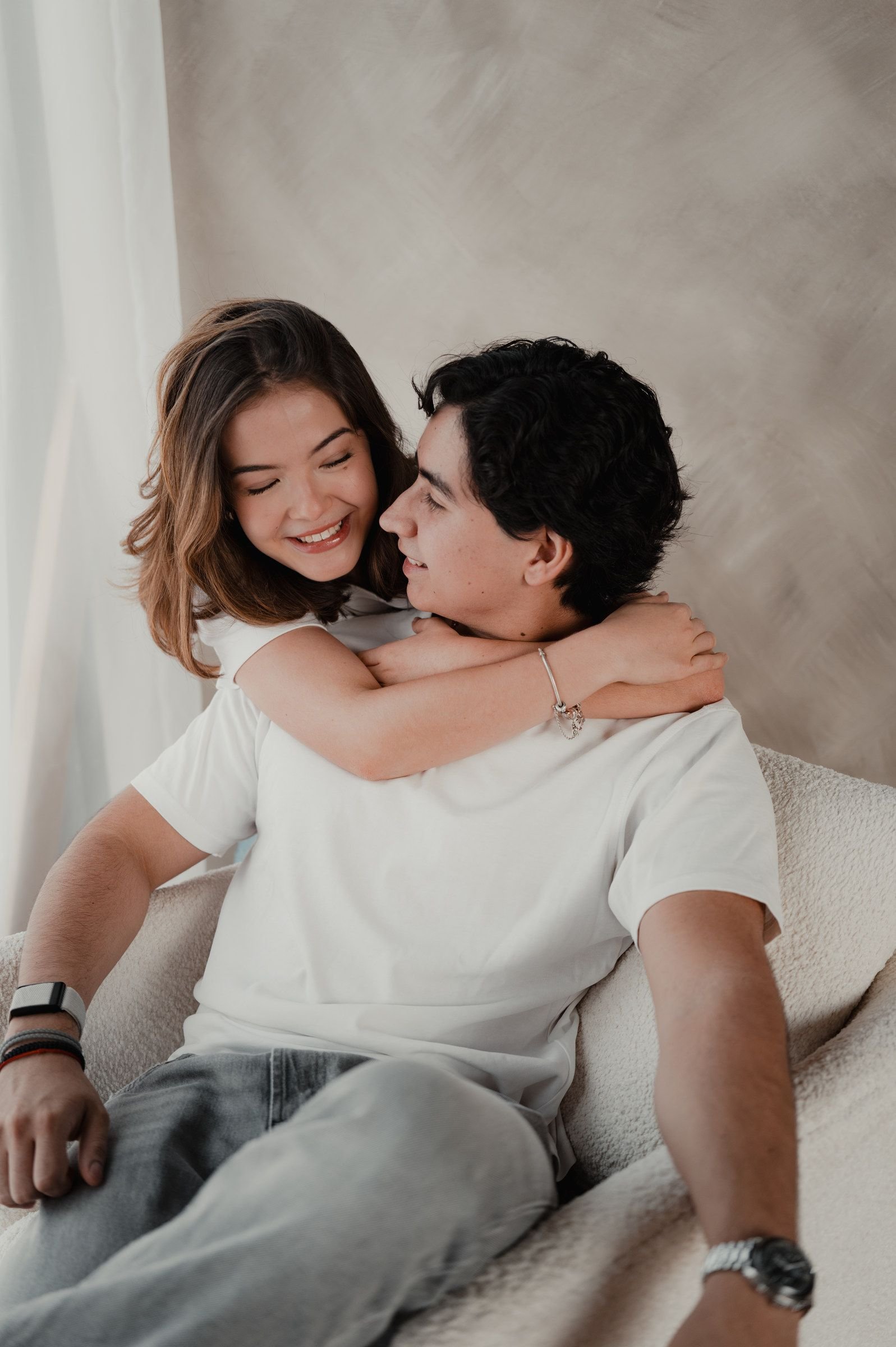 Ella lo abraza por el cuello y se ríe, sentados juntos en un sillón claro.