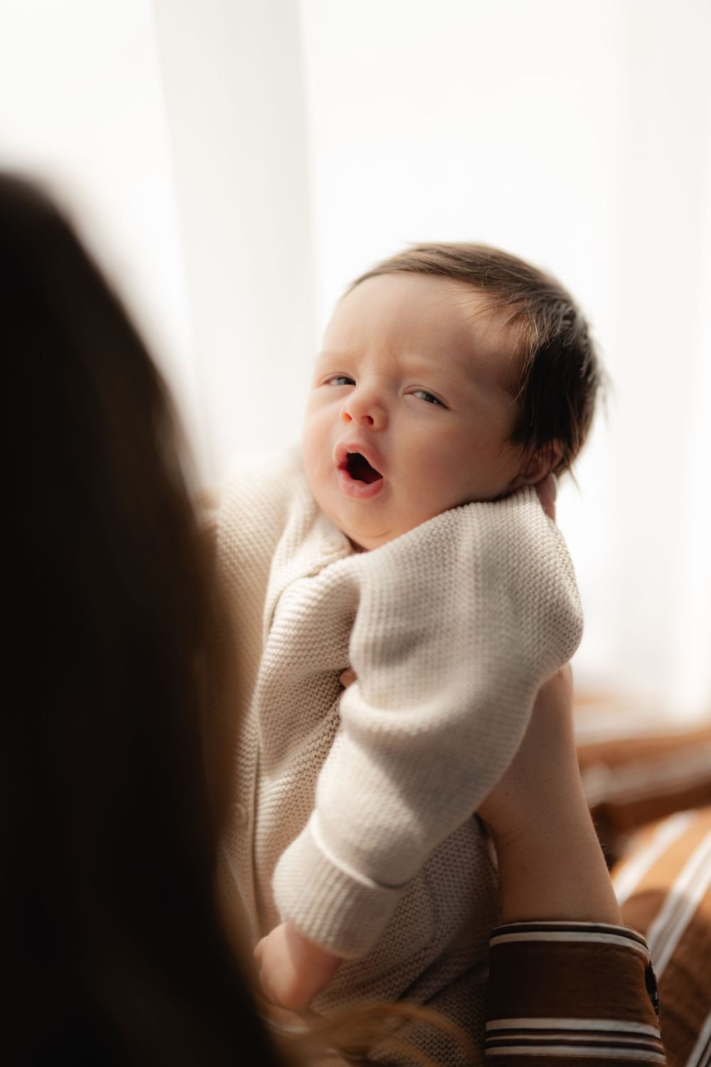 Un bebé recién nacido bosteza frente a una ventana luminosa, en brazos de su mamá.