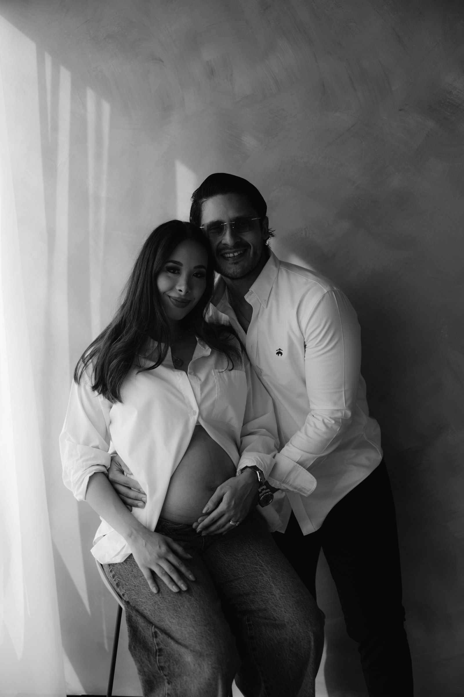 En blanco y negro, una pareja se abraza; ella lleva una camisa blanca abierta y él apoya las manos en su vientre.