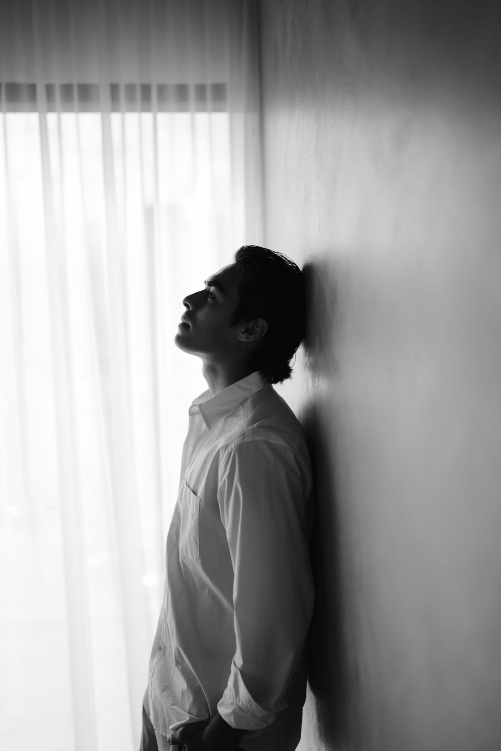 En blanco y negro, un hombre de perfil se recarga en la pared junto a una cortina.