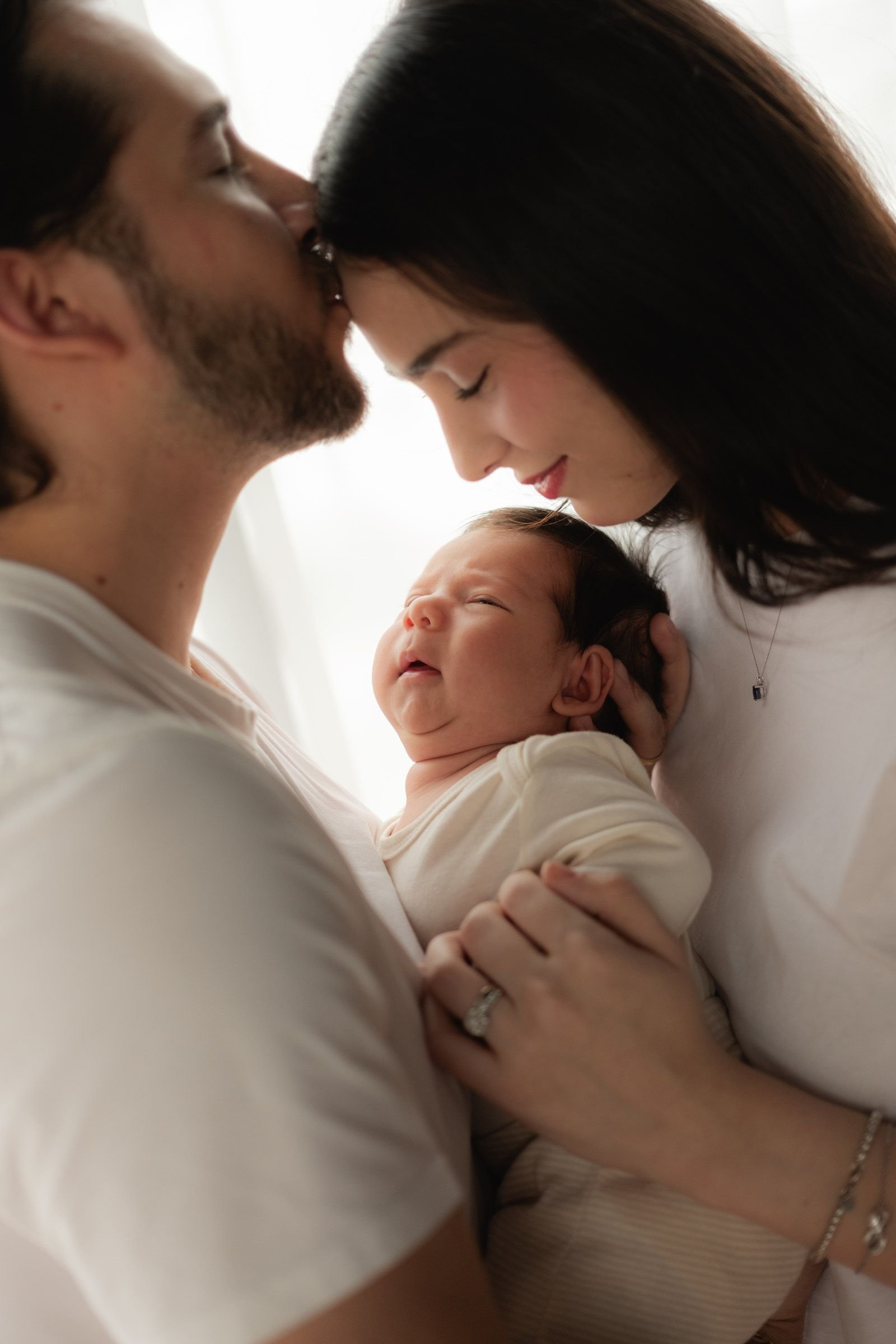 Mamá y papá se acercan frente con frente, con su bebé en medio, a contraluz de la ventana.