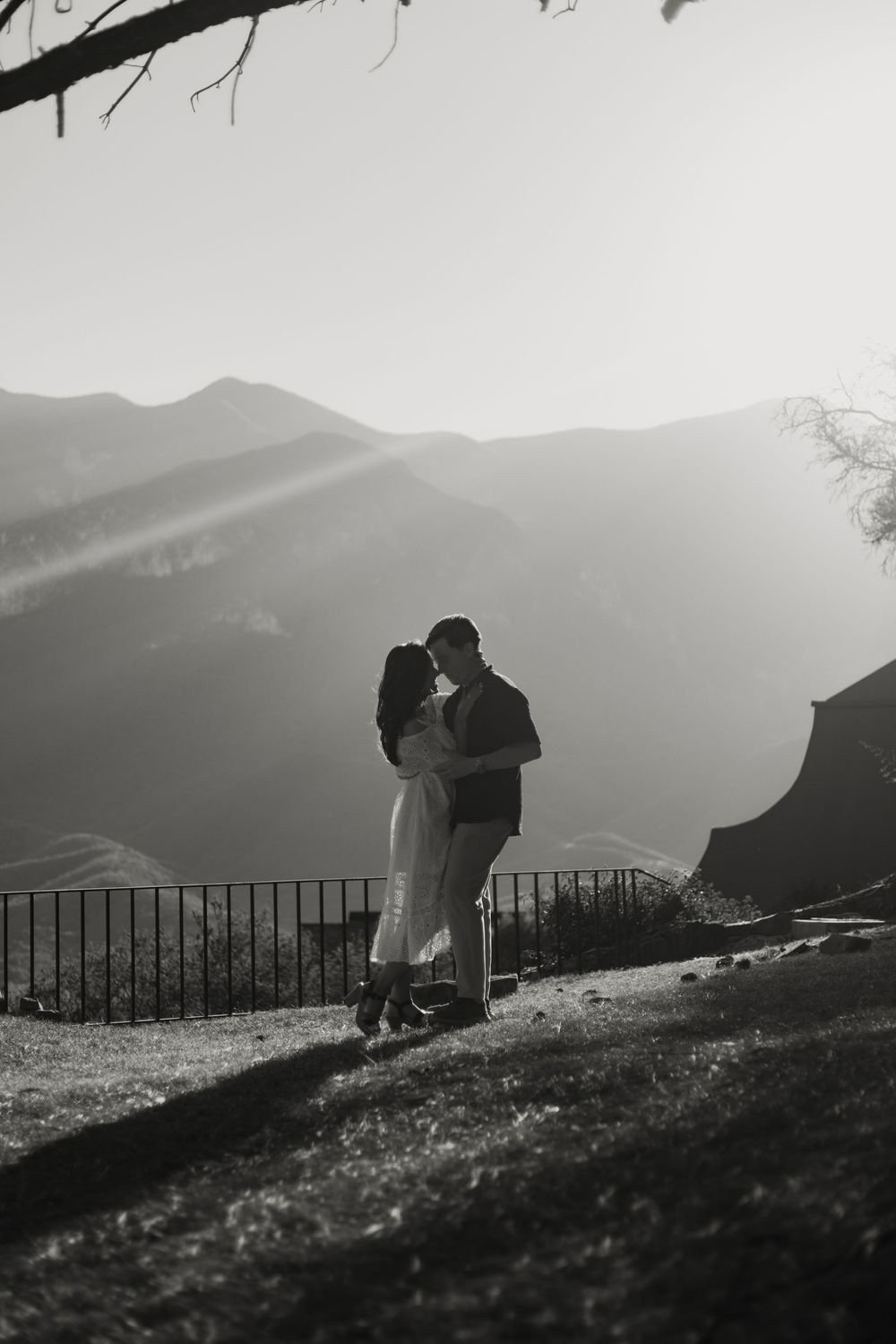 En blanco y negro, una pareja se abraza frente a las montañas, con el sol entre las nubes.