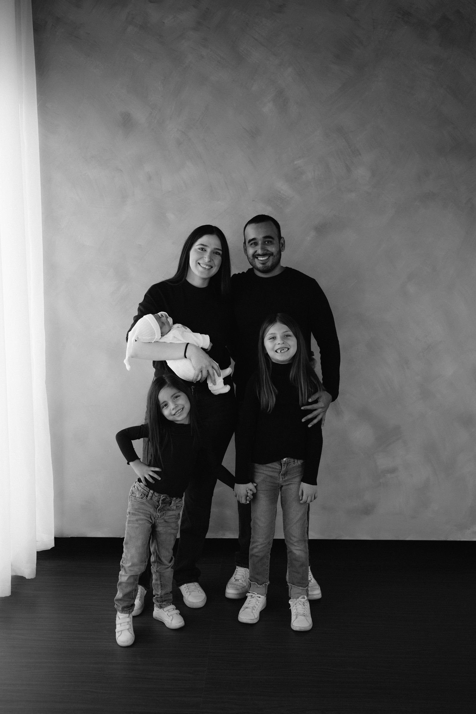 En blanco y negro, una familia posa de pie en el estudio, con los papás y sus hijas.