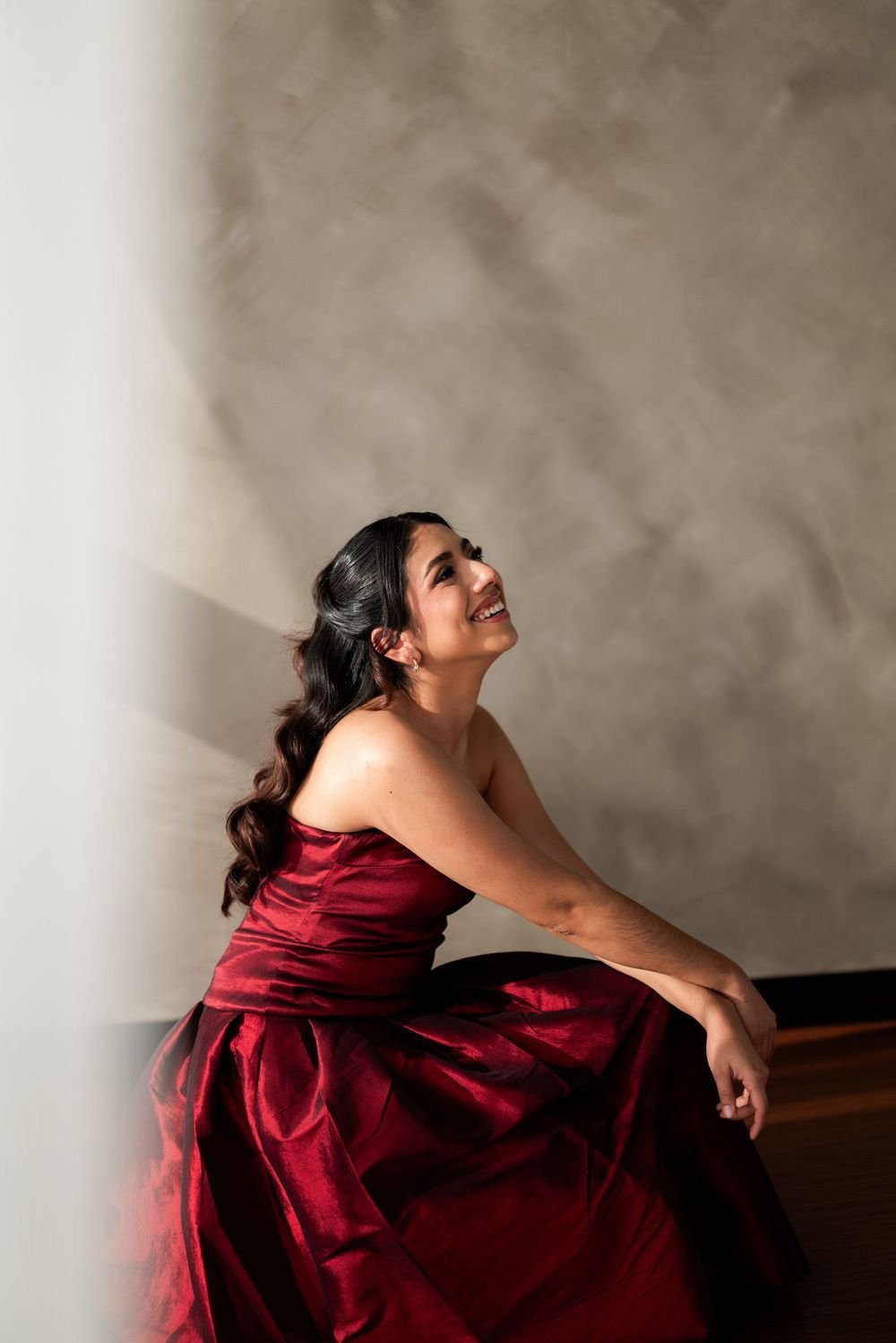 Una mujer con vestido rojo de satín sonríe mirando hacia arriba, sentada junto a la ventana.