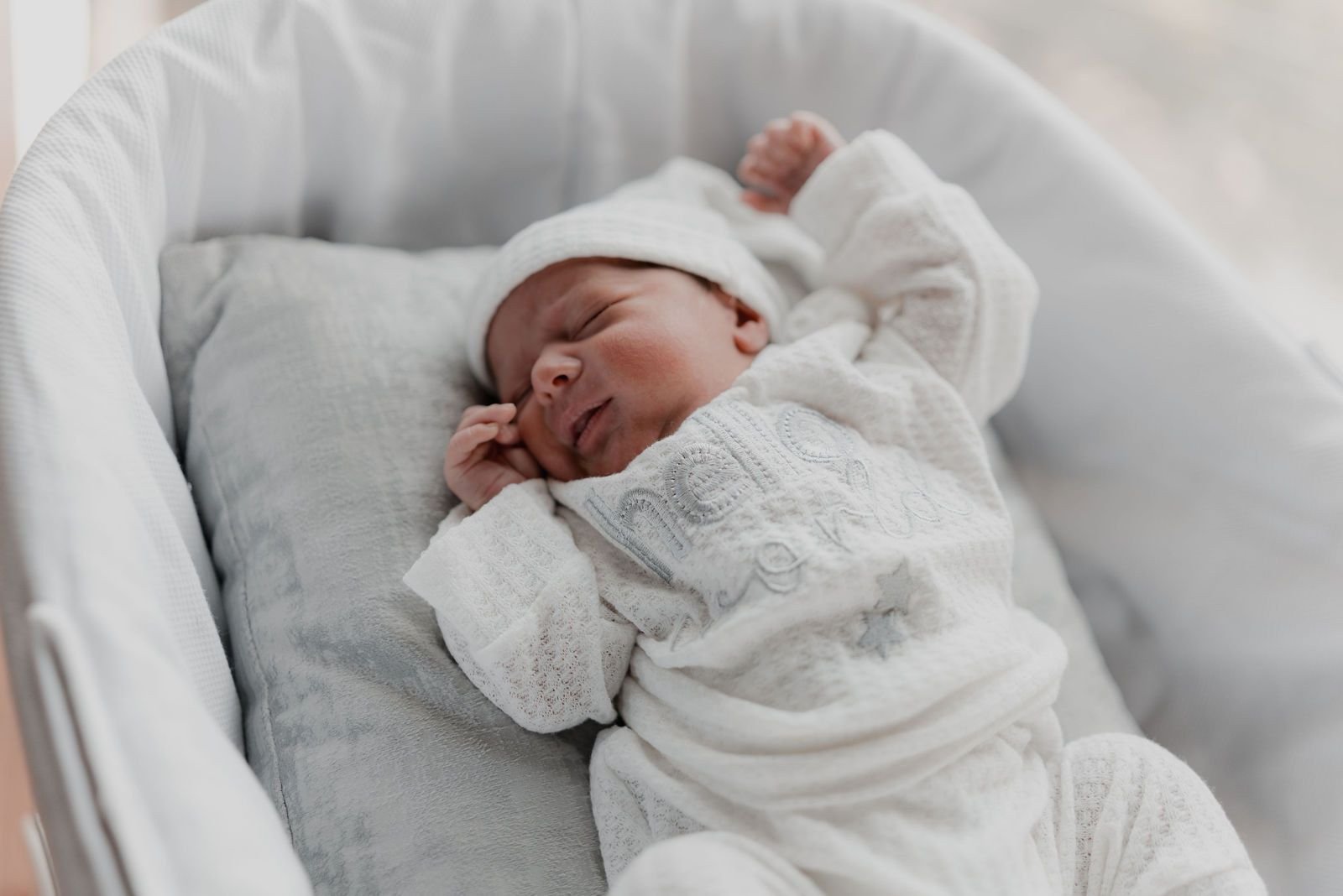 Un bebé recién nacido, con gorro y mameluco blancos, duerme en un moisés claro.