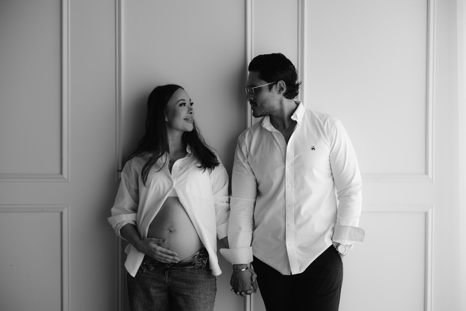 En blanco y negro, una pareja con camisas blancas se toma de la mano frente a una pared con molduras.